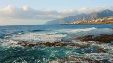 Atlantik Okyanusu 'nun boş kayalık volkanik plajı yumuşak sabah ışığında kırılan dalgaların üzerinde alçaktan uçuyor. Deniz köpüğü. Arka planda tatil köyü, şehir manzarası. Tenerife Kanarya Adaları İspanya Avrupa.