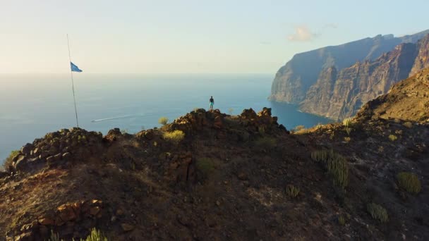 夕日の海の見える山頂に立つハイカー 旗が崖の上を飛んでいる 自然の風景 空中からの眺めテネリフェ島のギオスビーチにあるロス ギガンテスの崖 ロッキー形成 — ストック動画
