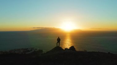 Hiking man in mountains watching sunset and horizon over beautiful ocean water landscape. Male hiker silhouette against colorful orange sky. Sun glare, sunburst. Shine.