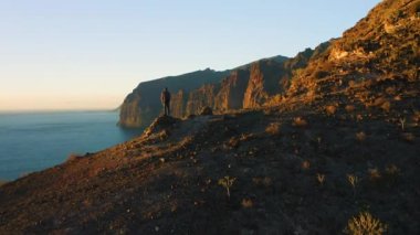 Man backpacker standing on mountain top with ocean view at sunset. Hiking healthy lifestyle concept. Natural landscape. Aerial view Los Gigantes cliffs over Guios beach on Tenerife Canay Islands Spain