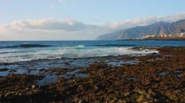 Okyanus dalgaları sahilin kayalık kıyılarına çarpıyor. Tuzlu deniz suyu köpüğü. Arka planda tatil köyü. Rocky volkanik Tenerife Adası. Şehir manzarası. İspanya Avrupa.