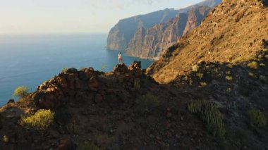 Hiker girl watching ocean view from rocky mountain top enjoying sunset nature in travel, wilderness trail. Lonely woman traveler in hiking contemplates landscape. Healthy lifestyle concept. Tenerife