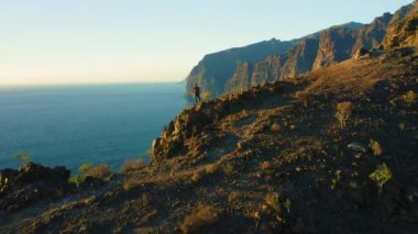 Rear view of man hiker stands on top of mountain cliff with ocean seascape. Success victory achievement concept. Famous tourictic spot Los Gigantes Tenerife Canary Islands Spain Europe.