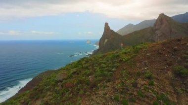 Hiker girl walking on the mountainside on edge of cliff with deep ocean and mountain landscape. Green coastline steep crags Roque de las Animas. Anaga Rural Park, Taganana in Tenerife Canary Islands