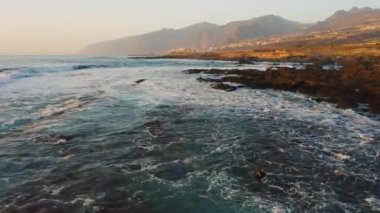 Rapid close-up drone movement over ocean water surface. Sea waves foam. Amazing scenery with Los Gigantes cliffs at warm sunset light. Fast high speed flight. Endless clear blue sky horizon.