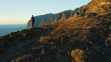 Rear view of man hiker celebrating climbing to the top of mountain cliff with ocean seascape. Success victory achievement concept. Famous tourictic spot Los Gigantes Tenerife Canary Islands Spain