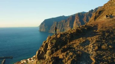 Man backpacker standing on top mountain cliff with rocky beach and ocean at sunset. Healthy lifestyle and travel concept. Aerial view of resort town Puerto de Santiago, Los Gigantes. Tenerife Canary
