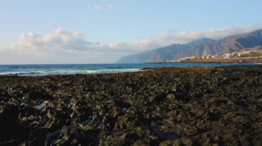 Foamy waves rolling towards rocky volcanic beach. Los Gigantes cliffs on the horizon. Blue cloudy sky. Drone fly close to water surface. Endless ocean seascape. Punta Juanita on Island Tenerife, Spain