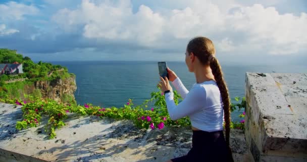 スマートフォンを使って岩の多いビーチでリラックスした女性 女の子は携帯電話で海の写真を撮る ウルワツ バリ島インドネシア 石灰岩の切り立った崖と紺碧のインドの水 泡の波 — ストック動画