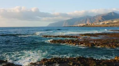 Atlantik Okyanusu 'nun boş kayalık volkanik plajı yumuşak sabah ışığında kırılan dalgaların üzerinde alçaktan uçuyor. Deniz köpüğü. Arka planda tatil köyü, şehir manzarası.