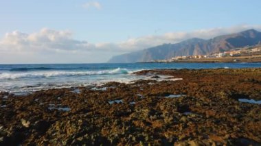 Köpüklü dalgalar kayalık volkanik plaja doğru yuvarlanıyor. Los Gigantes uçurumları ufukta görünüyor. Mavi bulutlu gökyüzü. İnsansız hava aracı su yüzeyine yakın uçar. Sonsuz okyanus manzarası.
