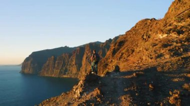 Erkek turist gün batımında okyanus manzaralı dağ tepesinde yürüyor. Yürüyüşçü, Tenerife 'de mavi gökyüzüne karşı Los Gigantes tepelerine tırmanışı kutluyor.