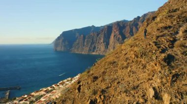 Sırt çantalı adam gün batımında okyanus kıyısında dağın tepesinde duruyor. Sırt çantalı turist manzarası derin deniz sularındaki kayalık kayalıkları küçümsüyor.