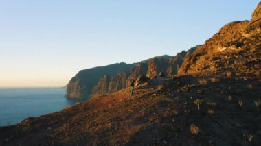 Erkek turist gün batımında okyanus manzaralı dağ tepesinde yürüyor. Yürüyüşçü, Tenerife 'de mavi gökyüzüne karşı Los Gigantes tepelerine tırmanışı kutluyor.