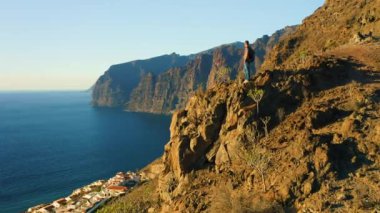 Sırt çantalı bir adam, gün batımında kayalık sahil ve okyanusla birlikte dağın tepesinde duruyor. Sağlıklı yaşam tarzı ve seyahat konsepti. Puerto de Santiago, Los Gigantes tatil köyünün hava manzarası. Tenerife.