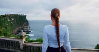 Kadınlar Uluwatu Hindu Tapınağı Pura Bali Adası Endonezya 'daki kayalık okyanus manzarasının tadını çıkarıyor. Turistik yerleri ziyaret ediyorum. Kızın arkadan görünüşü el kaldırıyor. Kireçtaşı sarp kayalıklar ve gök mavisi Hint Okyanusu suları