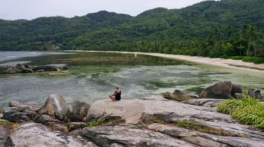 Cennet adasındaki egzotik tropik plajda granit kayalarda meditasyon yapan sarışın kızın hava manzarası. Meditasyon, sağlıklı yaşam ve uyum kavramı
