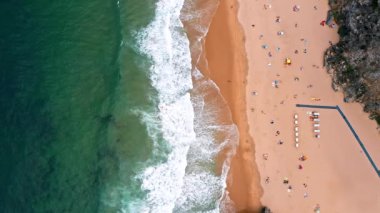 Praia da Adraga plajının 4K Aerial düzenli görüntüleri. Beyaz Atlantik Okyanusu dalgaları turistlerle kumsala doğru yuvarlanıyor. Sintra, Portekiz