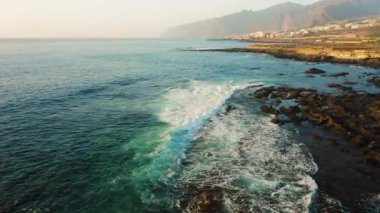 Su üzerinde alçaktan uçan dron, fırtınalı havada okyanus yüzeyinde. Deniz dalgaları köpürüyor. Kaya kıyısı. Arka planda Los Gigantes uçurumları var. Tenerife. Turkuaz denizde güzel bir manzara. Kanarya Adası.