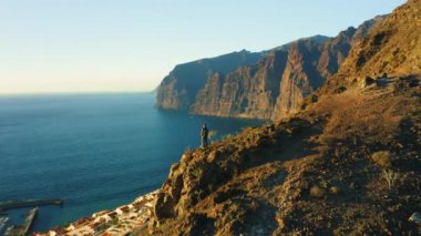 Sırt çantalı bir adam, gün batımında kayalık sahil ve okyanusla birlikte dağın tepesinde duruyor. Sağlıklı yaşam tarzı ve seyahat konsepti. Puerto de Santiago, Los Gigantes tatil köyünün hava manzarası. Tenerife.
