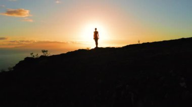Gün batımında dağın tepesinde bir kadın. Okyanus manzaralı dağlık arazide duran yürüyüşçü kız. Güneş ışığı bir kızın siluetini aydınlatır..