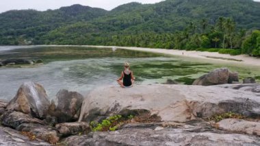 Yoga pozunda oturan ve cennet adasındaki egzotik tropik lagüne karşı granit kaya üzerinde meditasyon yapan sarışın kızın hava dairesi görüntüleri. Meditasyon, sağlıklı yaşam ve uyum kavramı