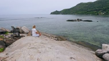 Sarışın, bronz tenli kızın yoga pozunda oturduğu ve granit kayalarda Cennet Adası 'ndaki egzotik tropik lagüne karşı meditasyon yaptığı hava manzarası. Meditasyon, sağlıklı yaşam ve uyum kavramı