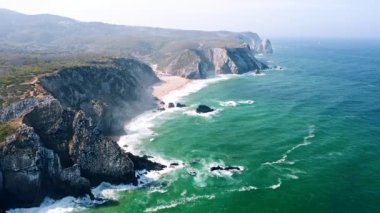 Aerial view of rocky cliff formation of Praia Grande beach, Portugal Atlantic coast. Cabo do Paco Lighthouse in Background.