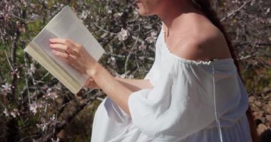 Woman reading a paper book in spring garden. Lady with book relaxing on nature outdoor. Enjoying vacation in flowering orchard.