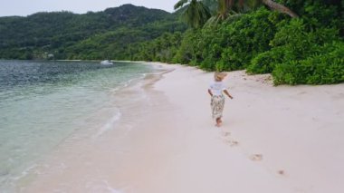 Kamera genç kadını yeşil yapraklı ve sığ lagünlü kumlu plajda koşarken izliyor. Güzel Mahe Adası 'ndaki bronzlaşmış kızın havadan 4K görüntüsü. Seyahat kavramı