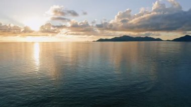 Gün batımının havadan 4K görüntüleri ve ufukta tropik ada silueti olan sakin okyanus görüntüleri. İnsansız hava aracını su yüzeyinin üstünde alçaktan uçuruyor.