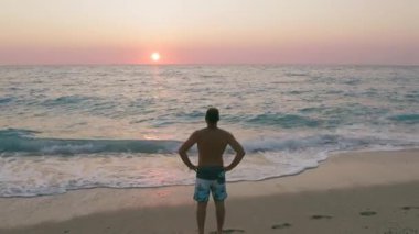 Sahil kenarındaki insan siluetinin statik görüntüsü. Gün batımında sakin dalgalarla deniz yüzeyinin tadını çıkarıyor. Turuncu gökyüzü parlıyor..