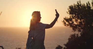 Kadın elinde telefonla gün batımında arka planda deniz olan bir selfie çekiyor. Yaz tatili hafta sonu