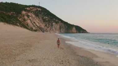 Alçak irtifa, Yunanistan 'ın İyon adası Lefkada' nın güzel Milos plajında eğlenen yetişkin bir kadının manzarasını takip ediyor. Gün batımı altın güneş ışığı ve el değmemiş dalgalar kıyı üzerindeki turkuaz mavi denizi kırıyor.