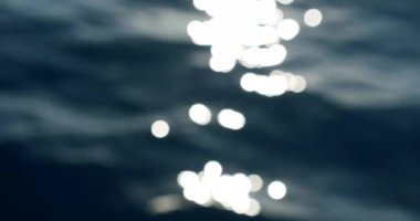 Bokeh güneşi su yüzeyine yansıyor. Güneş ışınları su yüzeyinde titreşiyor. Güneşin altında parıldayan küçük deniz dalgaları. Odak arkaplanından soyut bulanıklık. Yavaş çekim.