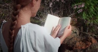 Kadın açık havada kitap okuyor. Baharın açtığı bahçede bilgi okuyan kız öğrencinin dikiz görüntüsü. Ağır çekimde yakın çekim.