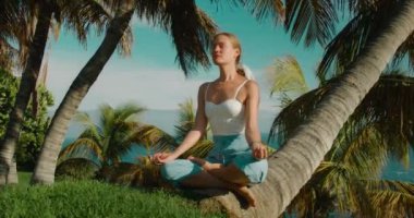 Tropik yeşil parkta yoga yapan bir kadın. Açık havada spor yapan yetişkin bir kızın yakın çekim görüntüleri. Sağlıklı yaşam tarzı kavramı. Arka planda palmiye ağaçları ve okyanus.