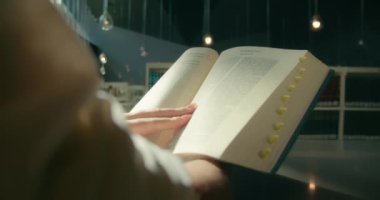 Kız öğrenci üniversite kütüphanesinde kutsal kitap eğitimi alan bir kitap okuyor. Sınavlara hazırlanıyorum. Arka planda kitap rafları olan yakın plan kadın elleri.