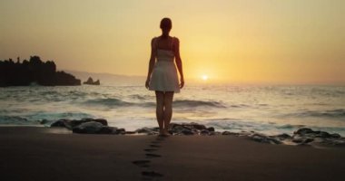 Plajda beyaz elbiseli bir kadın günbatımını seyrediyor ve kollarını kaldırıyor. Tatilde olan bir kadın silueti. Tatil ve seyahat konsepti. Pürüzsüz siyah kumda ayak izleri.
