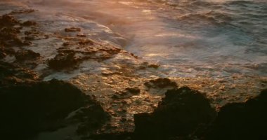 Altın gün batımında kayalık sahilde beyaz köpüklü okyanus dalgaları kırılıyor. Yakın çekim, volkanik sahildeki fırtınalı dalgalı deniz suyu. Sinematik görünüm.
