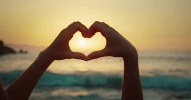 Gün batımında gün batımında denizde kalp şeklinde kadın elleri. Yavaş çekimde aşkın işareti. Tatildeki kadın silueti.