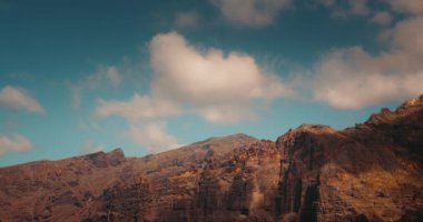 Tenerife kıyısındaki devasa volkanik dağların çorak tepelerinin görkemli manzarası. Mavi bulutlu güneşli gökyüzü. Sakin doğal manzara. Los Gigantes uçurumları.