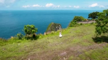 Nusa Penida Adası 'ndaki yeşil tropikal Kelingking plajından okyanus manzarası. Uçurumun kenarında yürüyen beyaz elbiseli turist kadın. Endonezya 'ya seyahat.