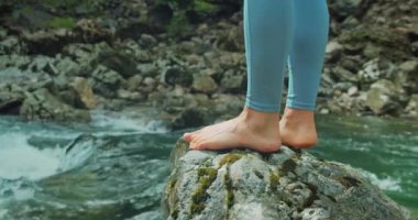 Nehir kıyısının kayalık kıyısında çıplak ayaklı bir kadın. Sağlıklı, güzel kadın bacakları. Yakın plan. Çıplak ayaklı bir kız dağlarda yürüyüş yaptıktan sonra dinleniyor dinlendirici bir dağ deresinin yanında..