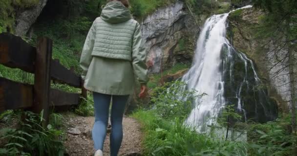 山の川沿いのハイキングトレイルを歩く女性と 雨の日の混雑した森で高い滝 アウトドアアクティビティと自然の中で旅行 — ストック動画