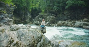 Kadın, hızlı bir nehrin yakınındaki taşların üzerinde lotus pozisyonunda meditasyon yapıyor. Dağ doğasında Zen yoga pratiği. Yürüyüşten sonra dinlenen bir kız. Sağlıklı yaşam tarzı.