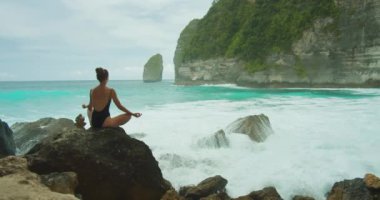 Denizin üzerindeki kayalık bir levrekte, siyah mayo giymiş bir kadın meditasyon yapıyor, engin okyanus önüne yayılıyor, arka planda dalgalar çarpıyor..