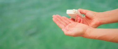 Kadın elinde beyaz bir kavanoz tutuyor kozmetik ürünü ve deniz suyu arka planında avucuna sıvı bırakıyor. Uzayı metin için kopyala. Cilt rutini, güzellik kremleri, temiz doğal. Deniz dostu.