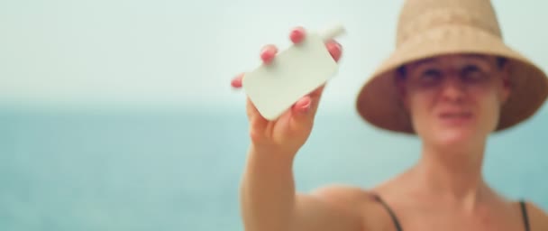 女性は使用前に日焼け止めを振ります 海の背景にスキンケアクリームの白いチューブを保持するビーチで水着の少女 女性はカメラに化粧品を展示しています テキストのスペースをコピーする — ストック動画
