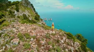 Capri adasının tepesindeki deniz manzarasına hayran bir kadın. Aşağıdaki körfezdeki berrak suları ve eğlence gemilerini çerçeveleyen yeşillikli kayalık kayalıklar. Yaz İtalya..